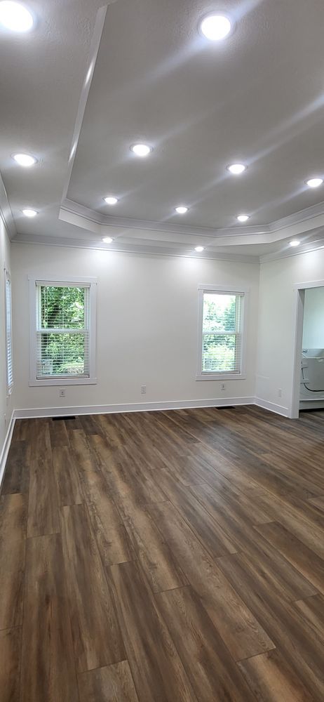 Empty living room with wood-look flooring, white walls, and recessed lighting. Windows offer a glimpse of greenery.