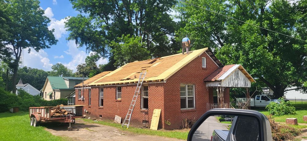 House under roof repair with worker on roof, ladder, and a trailer with debris in the yard.