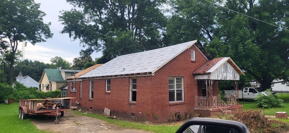 A brick house with a new roof and a small porch. A trailer and other houses are in the background.