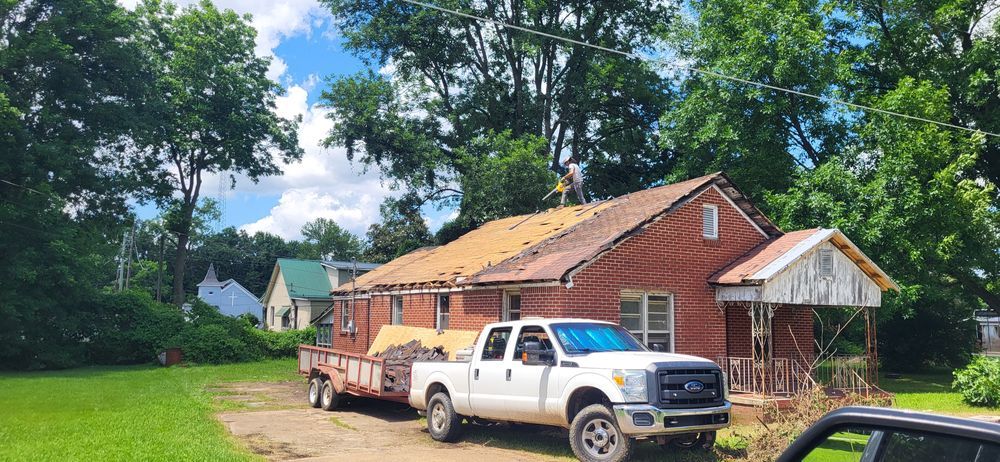 A white truck and trailer in front of a brick house with roof work being done. Trees and blue sky in the background.