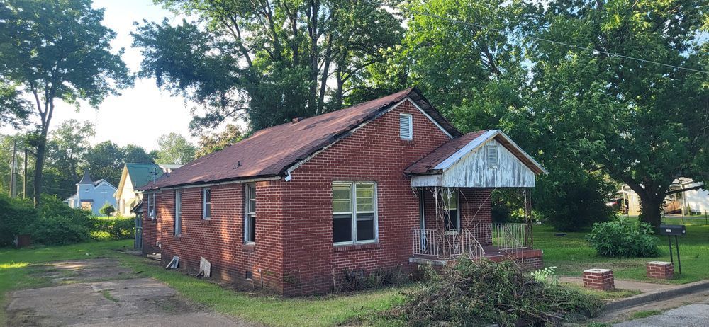 Red brick house with a porch and overgrown yard, set among trees and other houses.