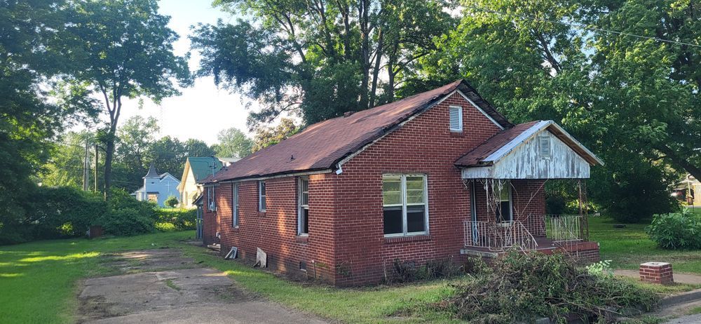 Red brick house with a weathered porch and roof, surrounded by trees and grass.
