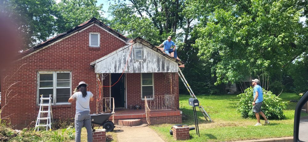 People working on a red brick house. One on the roof, others on the ground. Green trees surround the house.