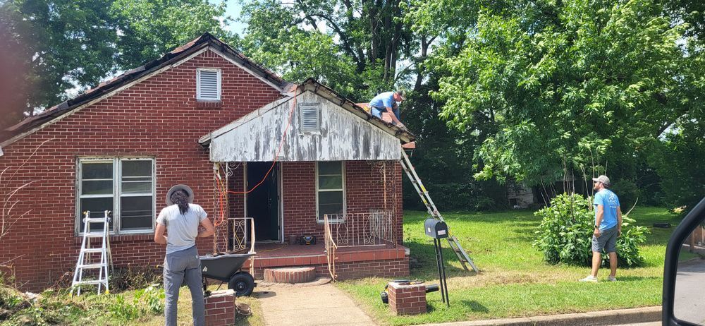 People working on the roof of a red brick house. One person is on a ladder. Green grass and trees are in the background.