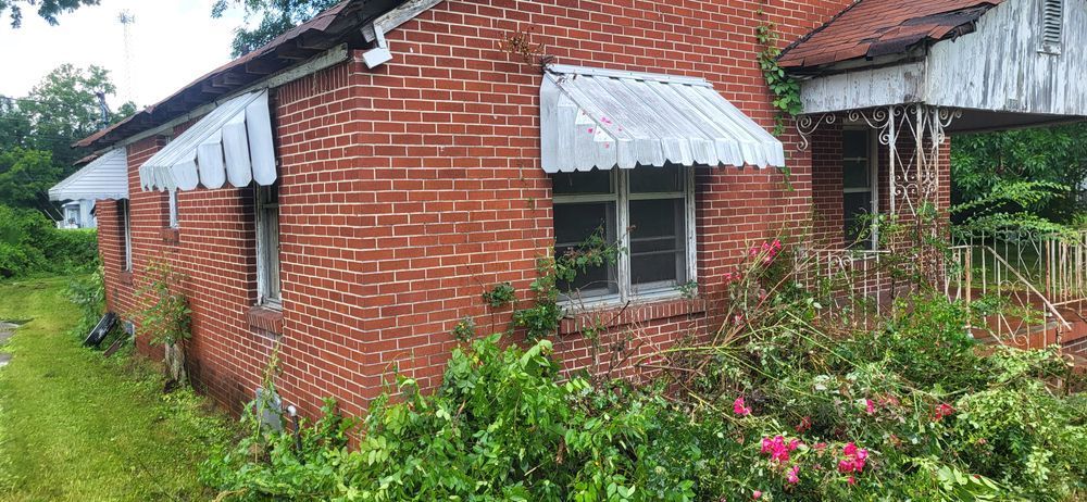 A weathered brick house with white awnings and overgrown plants.