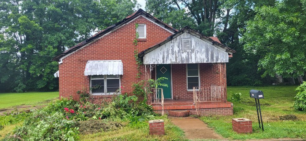 Brick house with green door, weathered porch, overgrown yard.