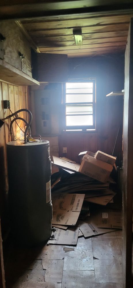 A small, cluttered room with a water heater, window, and pile of cardboard boxes.