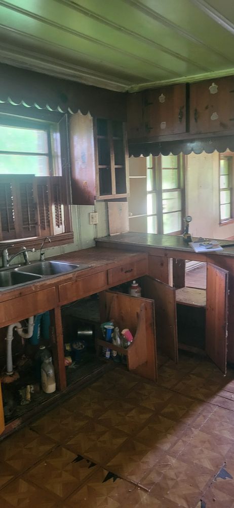 A dilapidated kitchen with wooden cabinets, counter, and a sink. The floor appears worn.