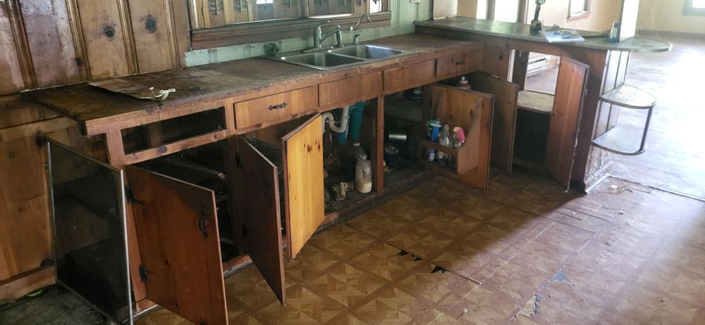 Old wooden kitchen with open cabinet doors, visible plumbing, and a worn countertop.