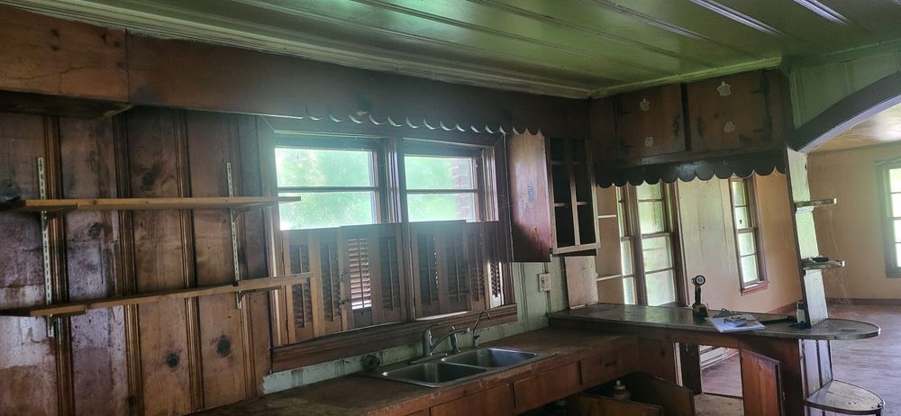 Interior of a dilapidated kitchen with exposed wooden walls and cabinets. A sink and countertop are visible.