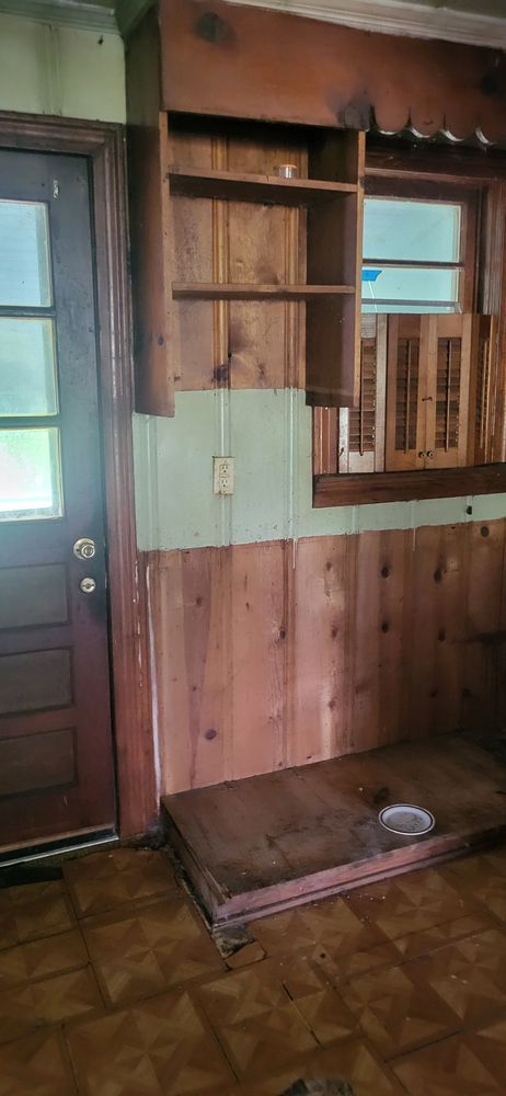 Interior view of a kitchen with wooden paneling, shelves, and a raised wooden platform. Brown and green tones.