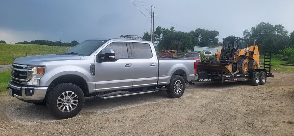Silver pickup truck towing an orange and black construction vehicle on a trailer. Gravel road, trees in the background.