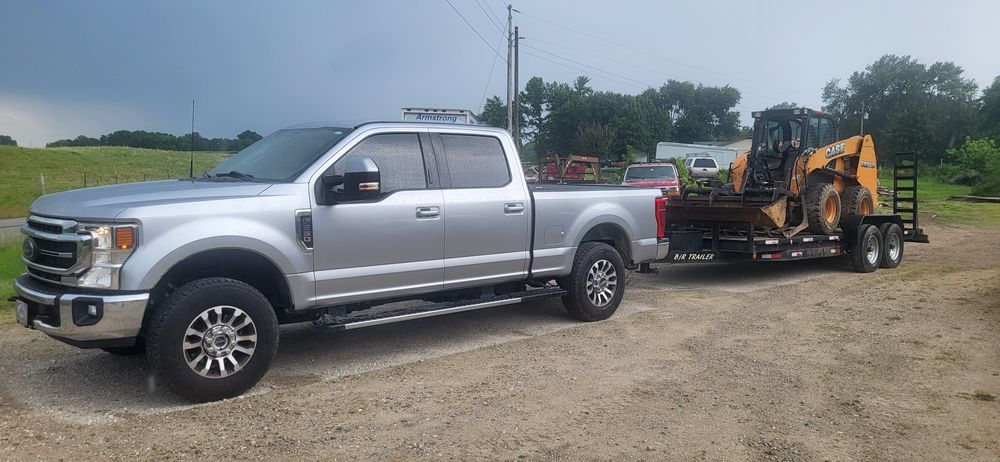 Silver pickup truck towing a trailer with a Case backhoe on a gravel driveway.