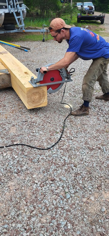 Man using a circular saw to cut a wooden beam outdoors. Gravel ground, blue shirt, brown hat.
