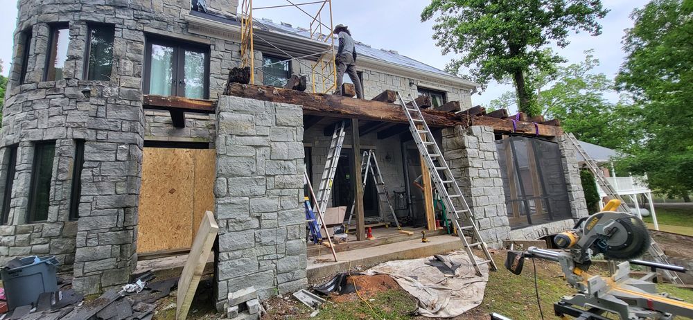 Exterior of a stone house under construction; a worker stands on the roof with ladders and tools.