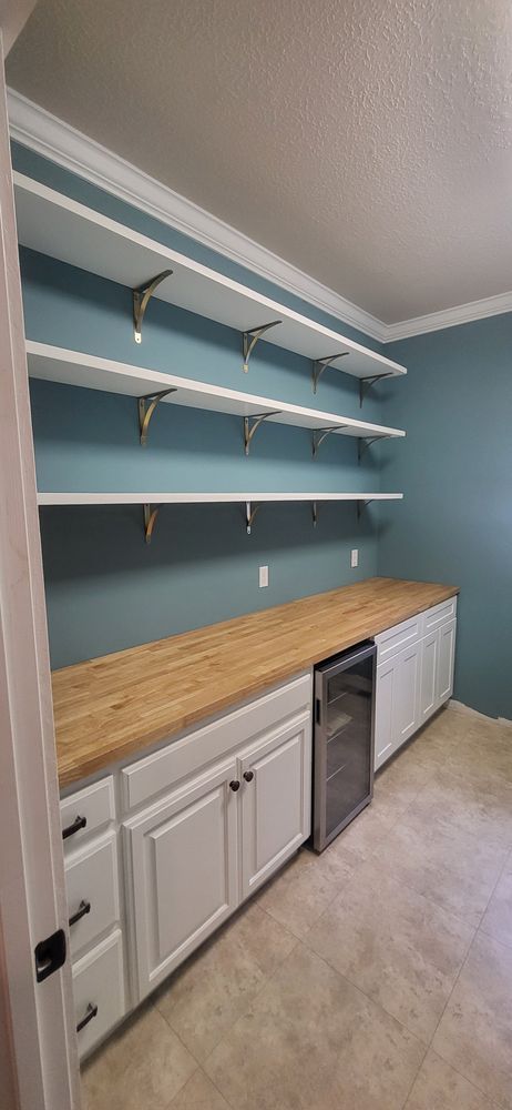 A pantry with white cabinets, wooden countertop, and shelves. A wine fridge is visible. Blue walls.