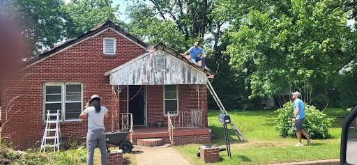People working on the roof of a red brick house. One person is on a ladder. Another is on the lawn.