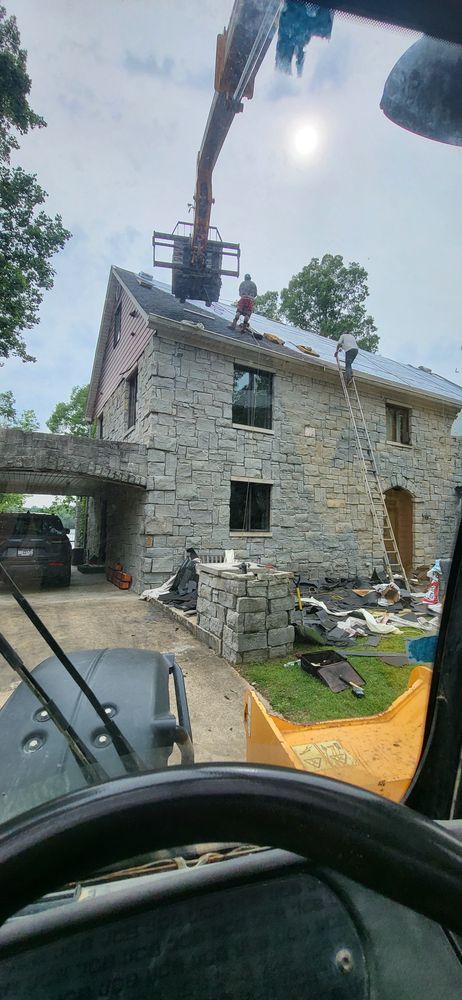 A house with a stone exterior has its roof worked on by construction workers. A crane is also present.