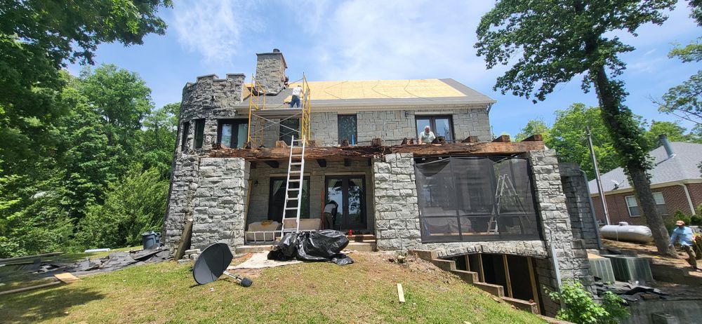 House under construction; stone exterior, exposed roof. Tall ladder against the structure. Overcast.