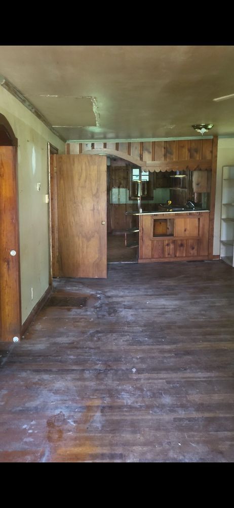 An empty room with a wooden floor and kitchen area visible through a doorway.