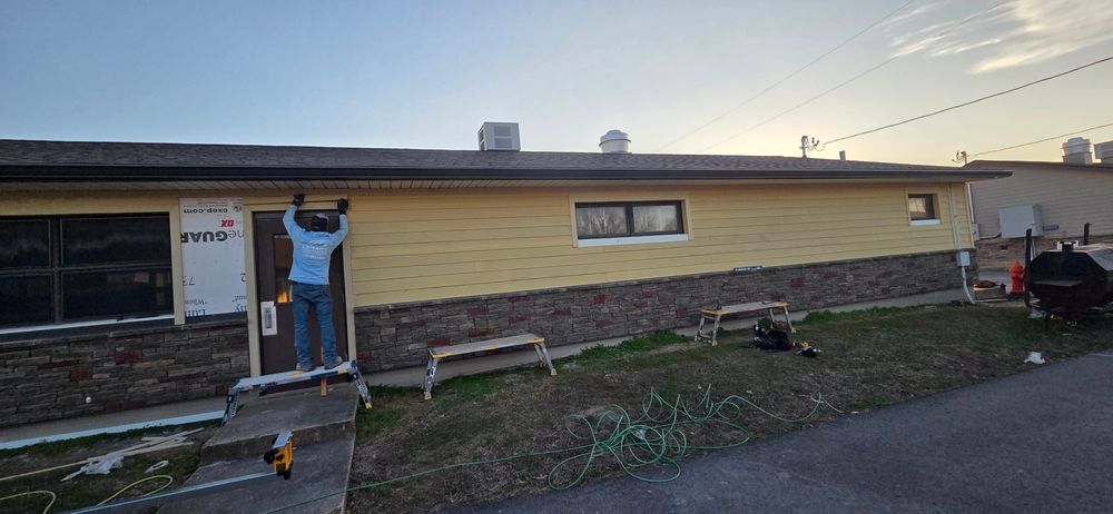 Person works on siding of a house with yellow siding and a stone veneer.