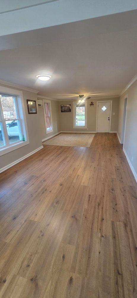Empty living room with wood-look flooring, neutral walls, white trim, and a white door at the end.