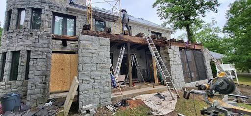 Construction work on a two-story stone house. A worker is on the roof. Ladders and tools are visible.