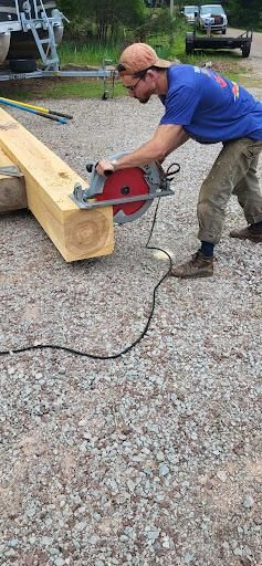 Man using a circular saw to cut a wooden beam outdoors. Gravel ground, blue shirt, brown cap.