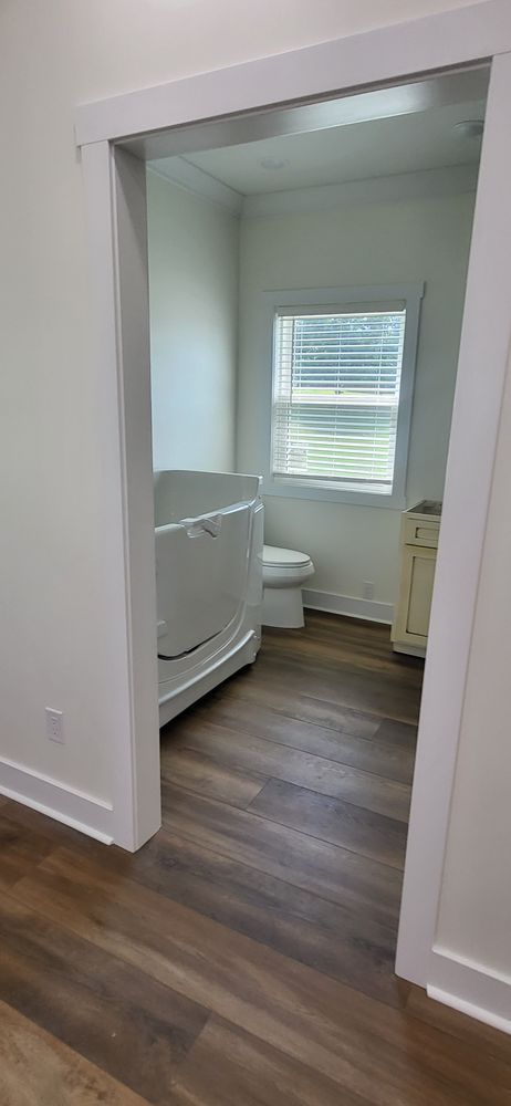 Bathroom with a walk-in tub and toilet visible through a doorway with wood-look flooring and white walls.