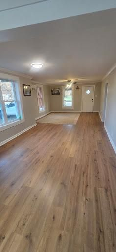 Empty living room with wood-look flooring, neutral walls, and a white front door. Sunlight streams through the windows.