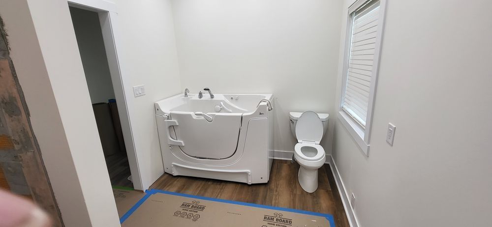 Bathroom with a walk-in tub and toilet. The walls are white with a window. The floor is wood.