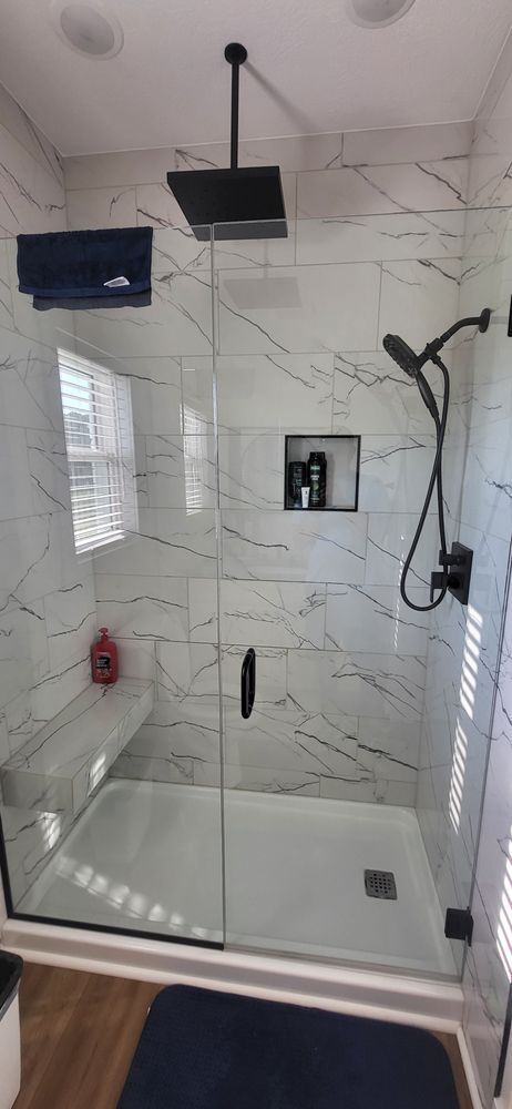 A modern shower with white marble-like tiles, a black showerhead, and a glass door.