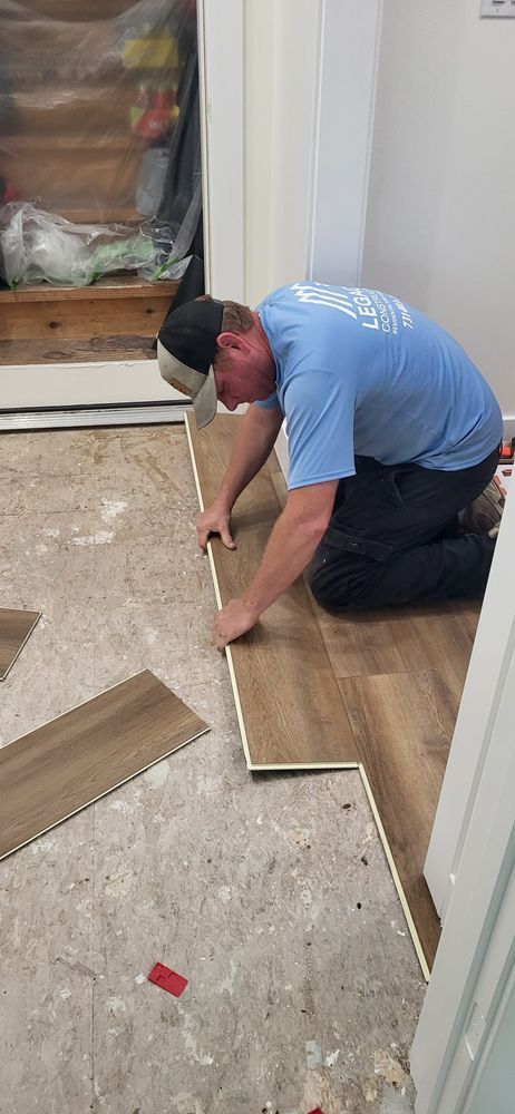 Person kneeling, installing wood-look flooring. Indoors, near a doorway. Light blue shirt, black pants, baseball cap.
