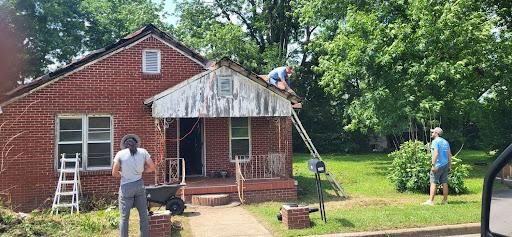 People working on a house with a red brick exterior, one on a ladder. Green lawn and trees.