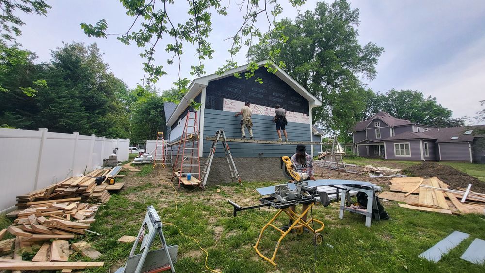 Construction workers siding a house, blue siding, black roof shingles. Yard with tools, lumber, and fencing.