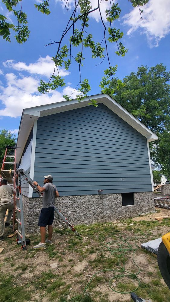 Workers install blue siding on a building with a stone foundation under a blue sky.