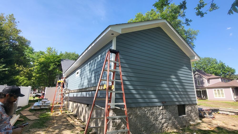 Construction on a house with gray siding and stone foundation. A person works near an orange ladder on a sunny day.
