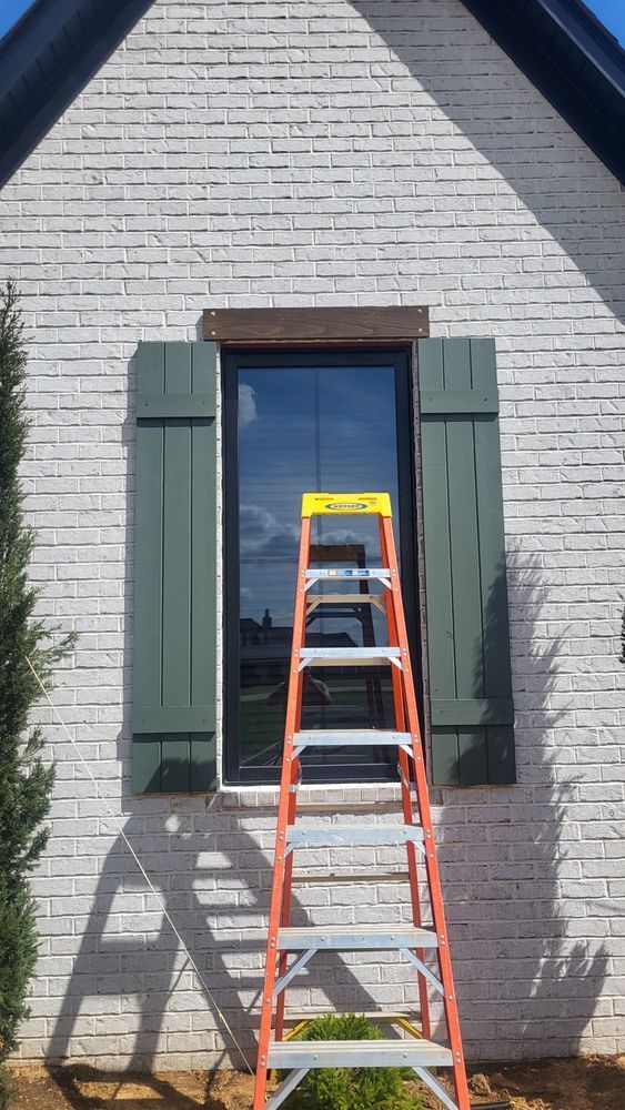 Ladder leaning against a house with green shutters and brick wall.