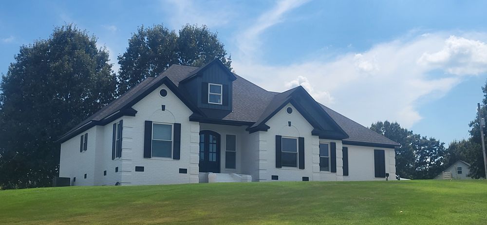 White house with black shutters, roof, and door trim, set on a green lawn against a blue sky.