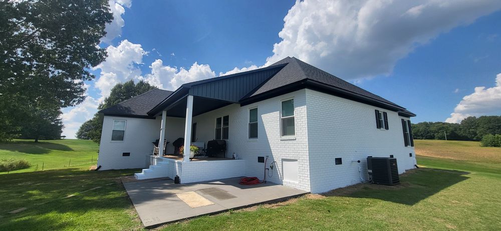 White house with black roof and a porch, on a green lawn, under a blue sky with clouds.