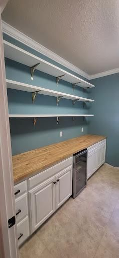 A kitchen pantry with white shelves, cabinets, and a wood countertop. Wine fridge and blue-green walls.