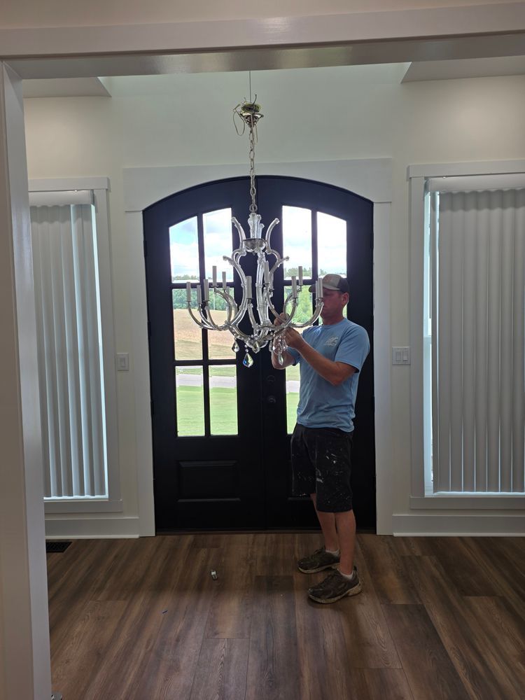 Person installing a chandelier in a doorway. Interior with wood flooring, black door, and white trim.