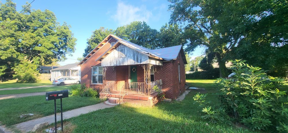 Brick house with a small porch, green door, and overgrown yard.  A mailbox is in the foreground.