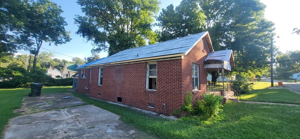 Brick house with a metal roof. A small porch is attached. Trees and grass in the yard.