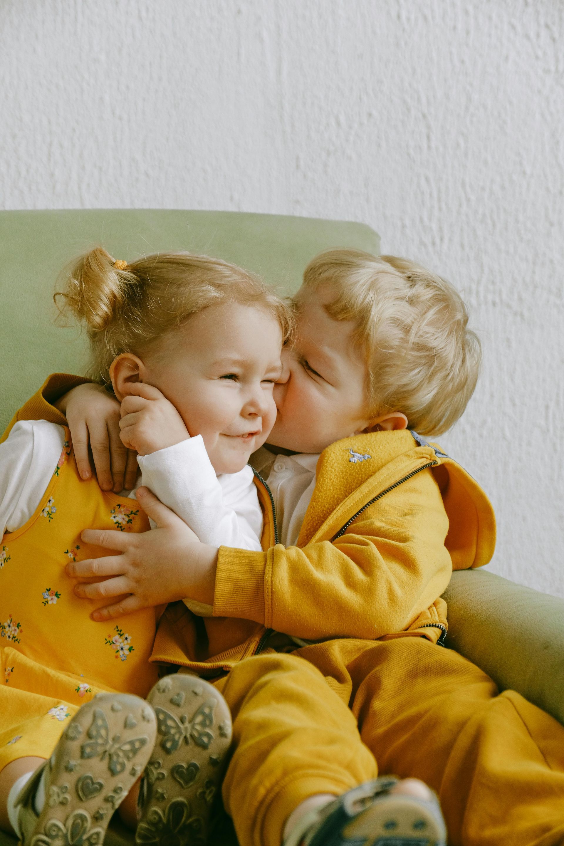 Boy in yellow kisses girl's cheek; both in yellow outfits, seated on a green chair.