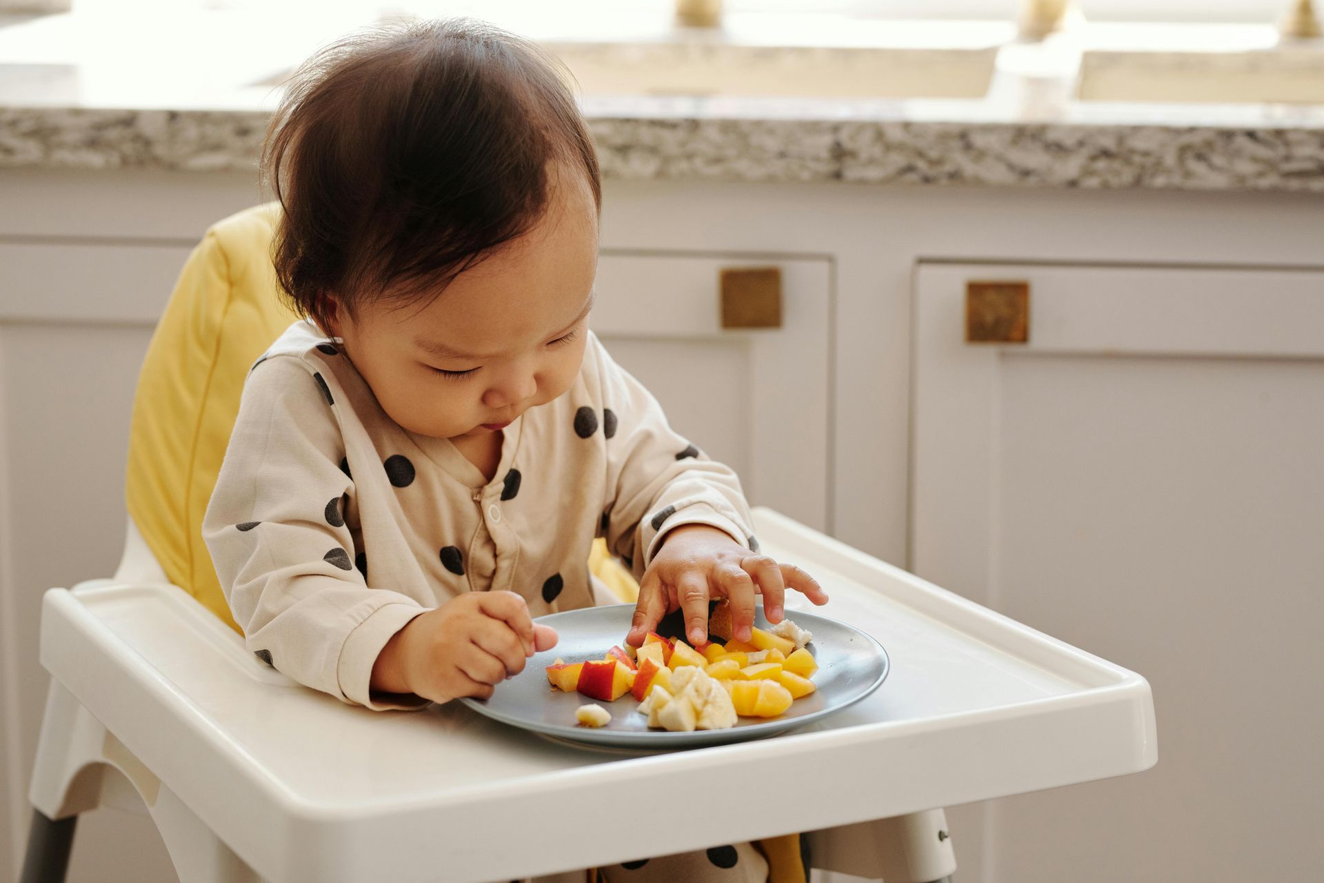 Baby in a high chair eating from a plate, leaning forward in a kitchen.