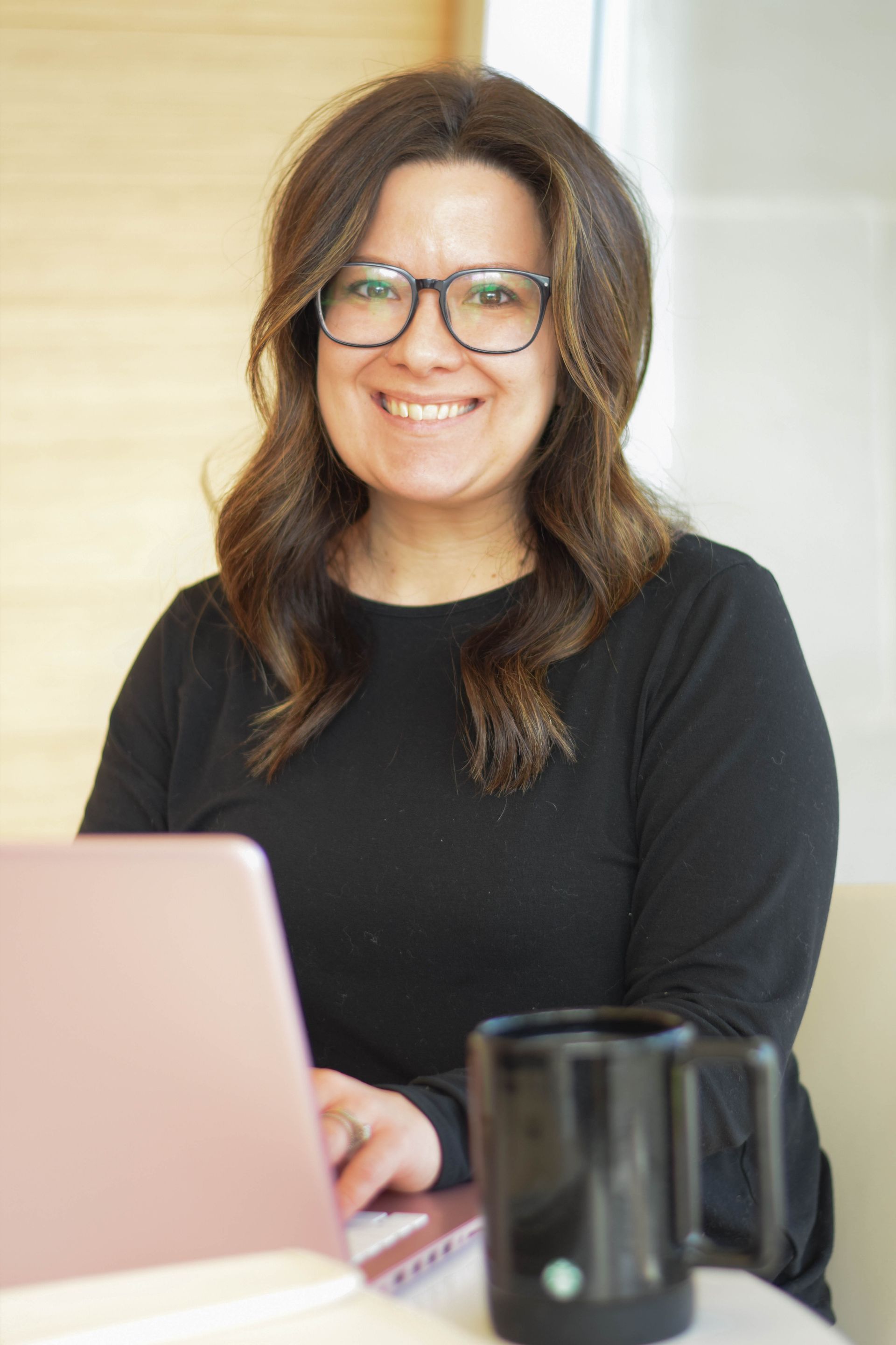 Woman with glasses smiles while working on a pink laptop, with a black mug nearby.