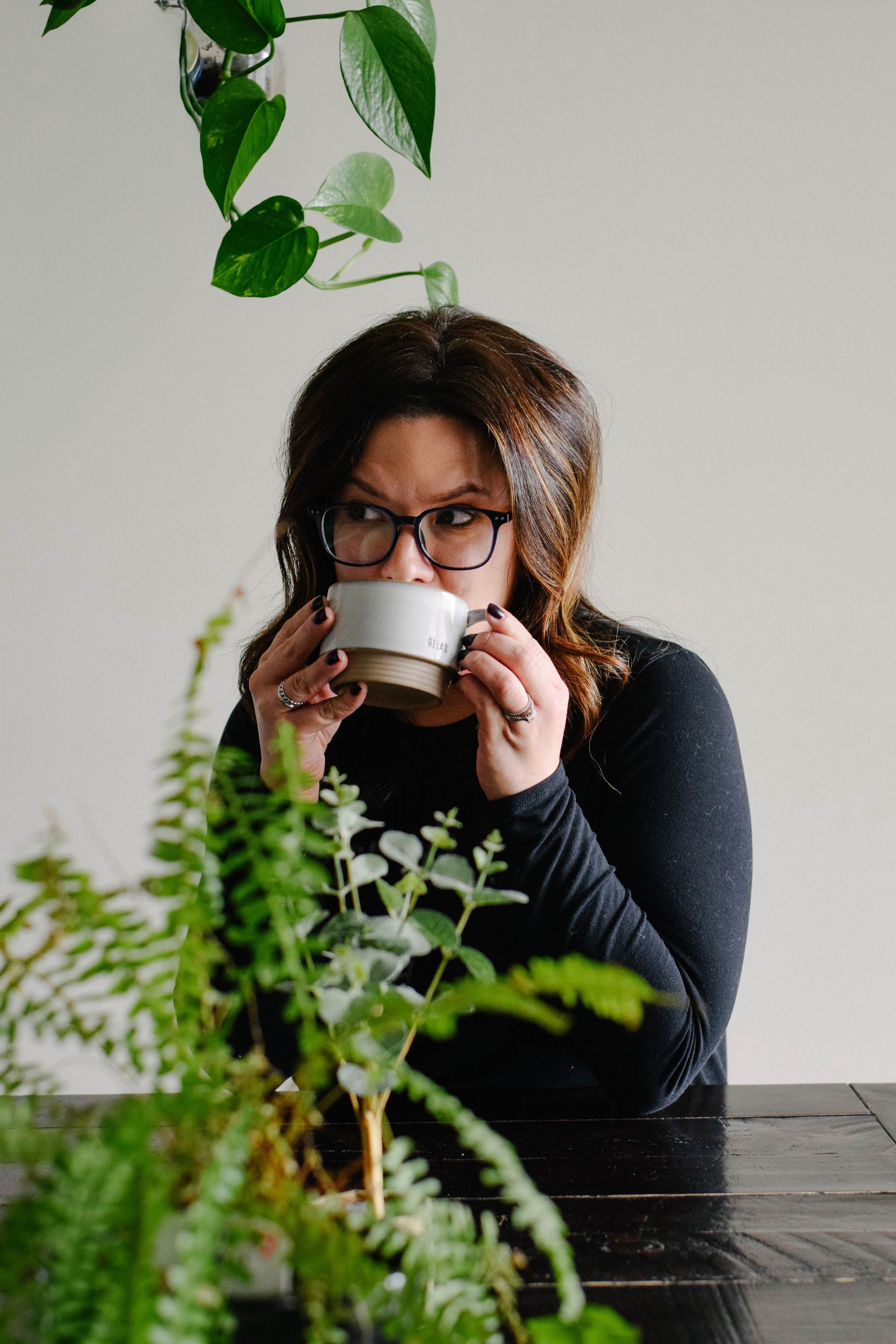 Woman sipping from a mug, wearing glasses, indoors with plants and a white wall.