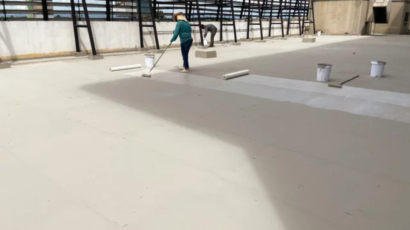 Person applying light-colored coating to a large, empty concrete surface outdoors with rollers and paint buckets.
