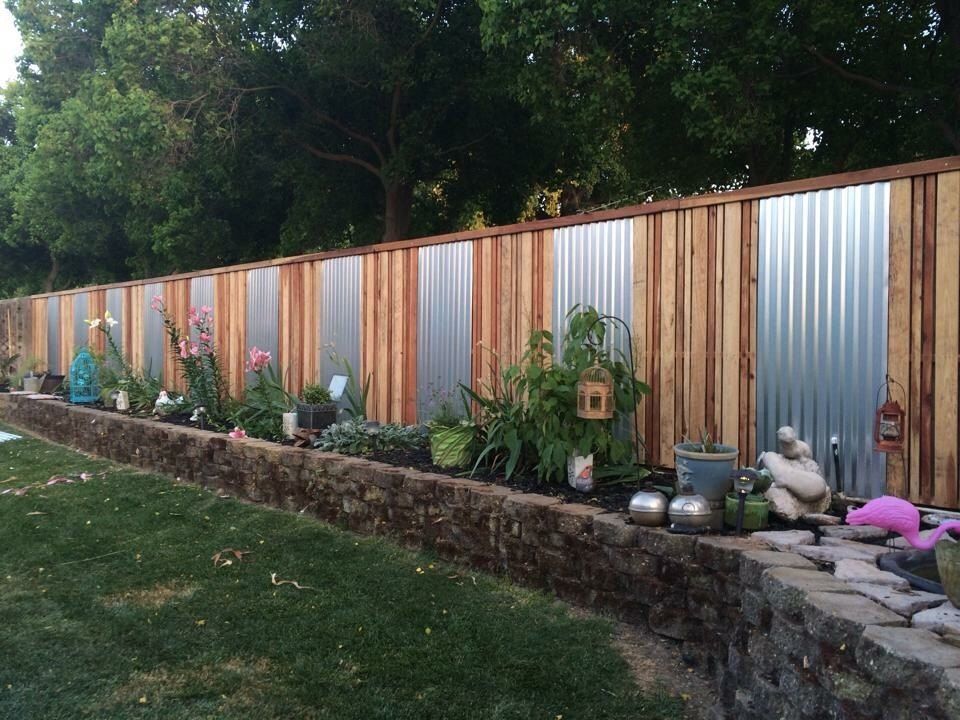 Fence with alternating wood and corrugated metal panels, bordering a stone-lined garden bed with flowers and greenery.